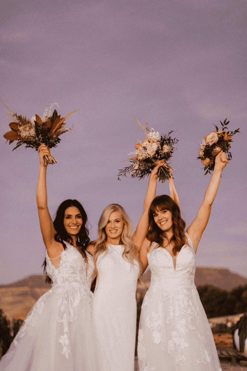 Three brides in white dresses holding flower bouquets, smiling and celebrating against a scenic backdrop.