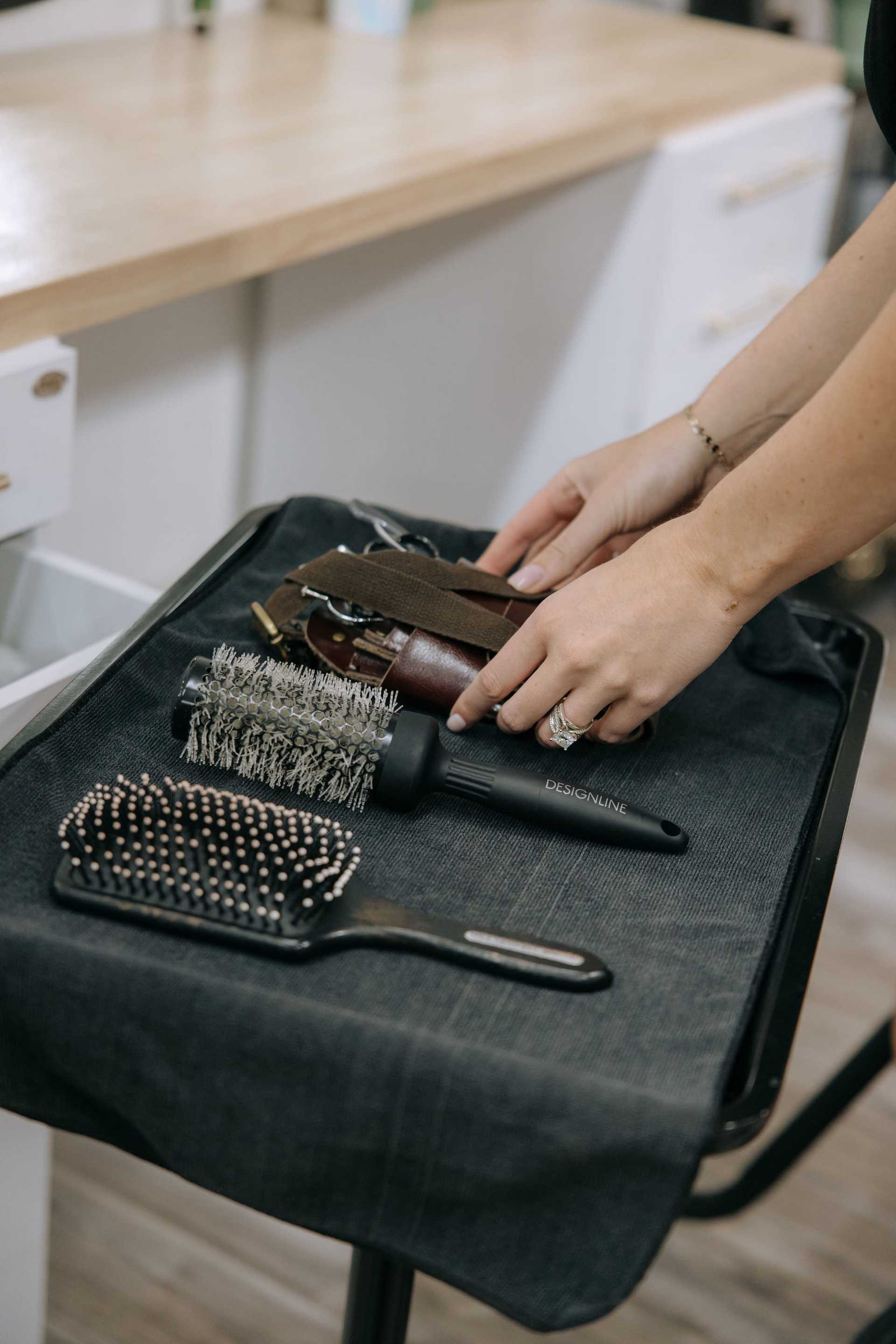 Hands organizing hair styling tools including a brush, a roller brush, and a leather pouch.