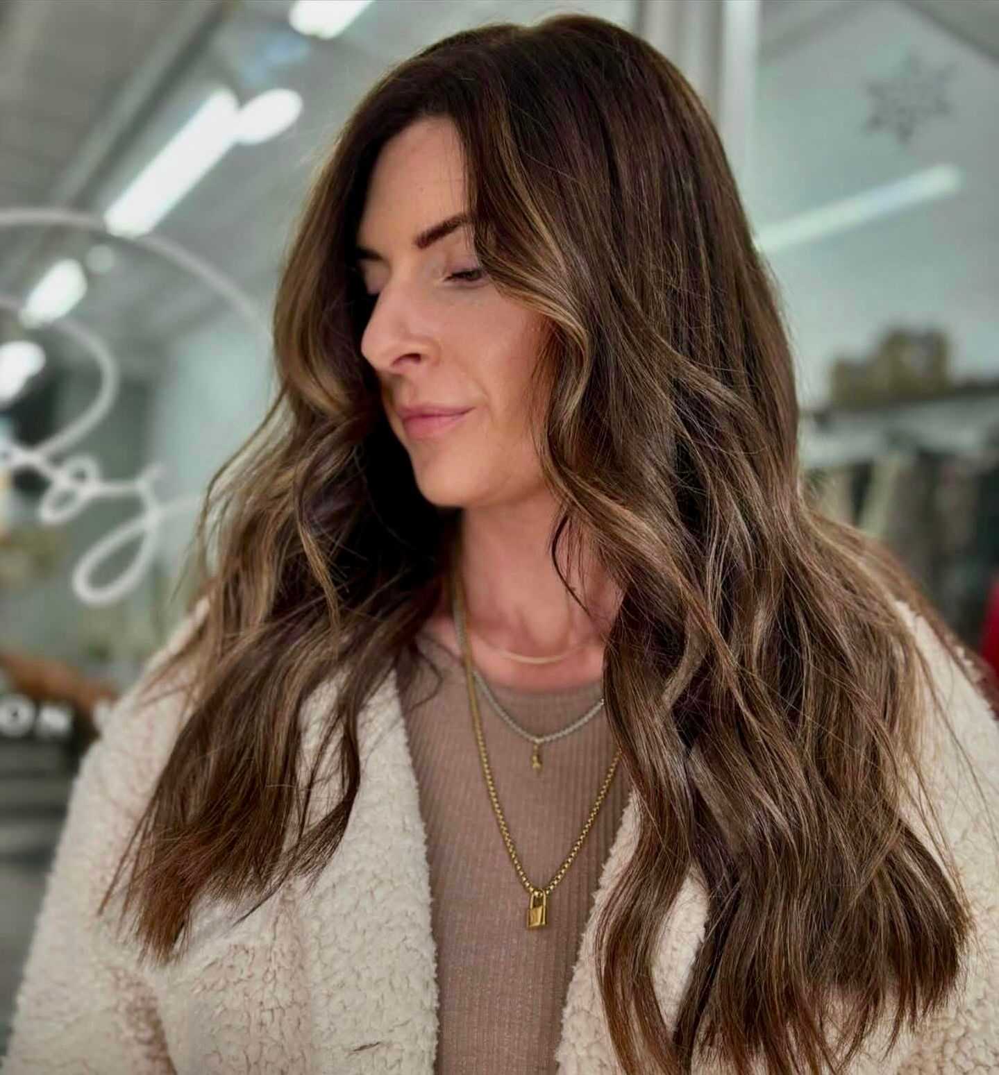 Woman with long wavy hair in a cozy jacket, standing indoors.