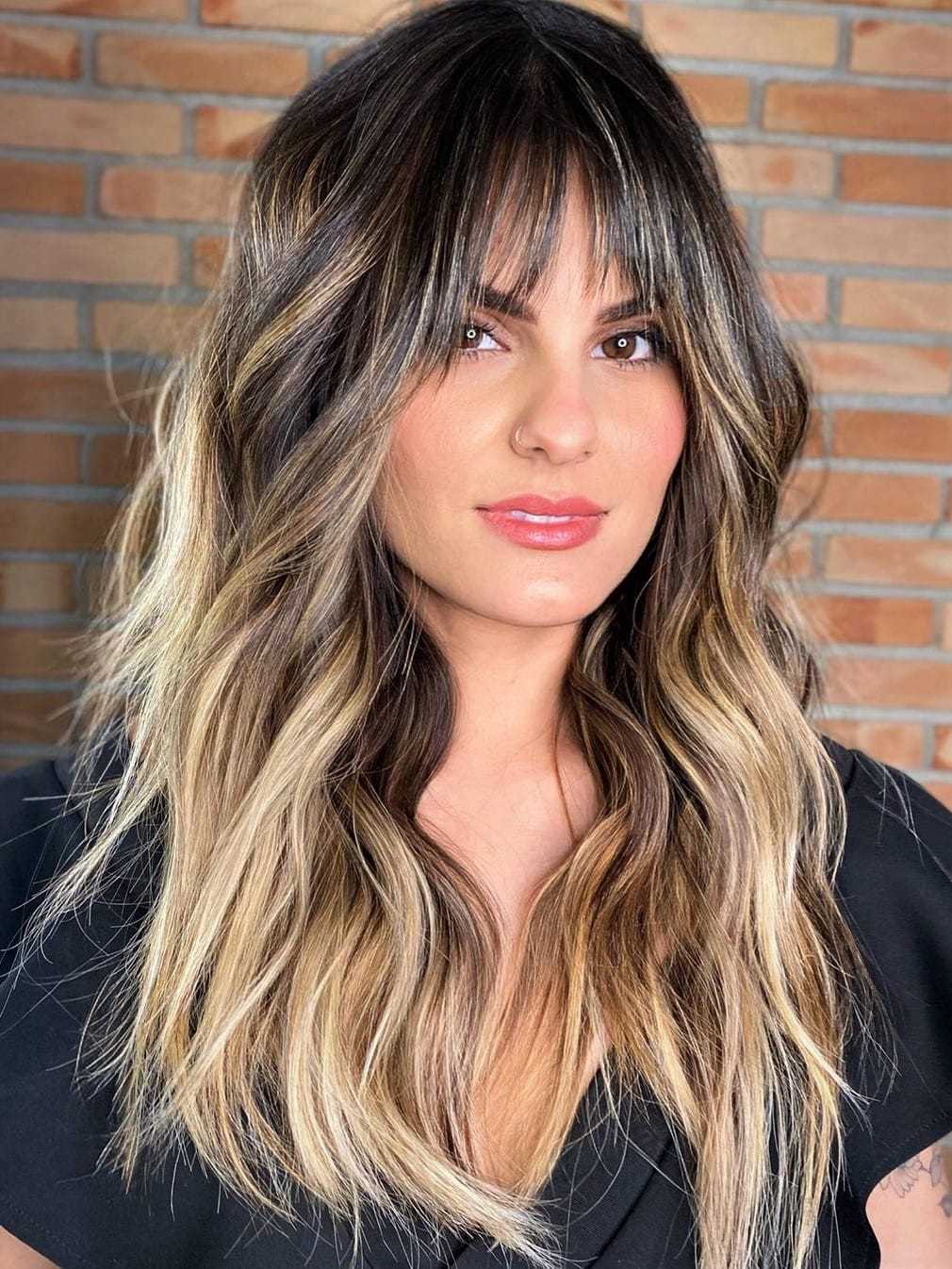 Woman with wavy balayage hair and bangs in front of a brick wall.