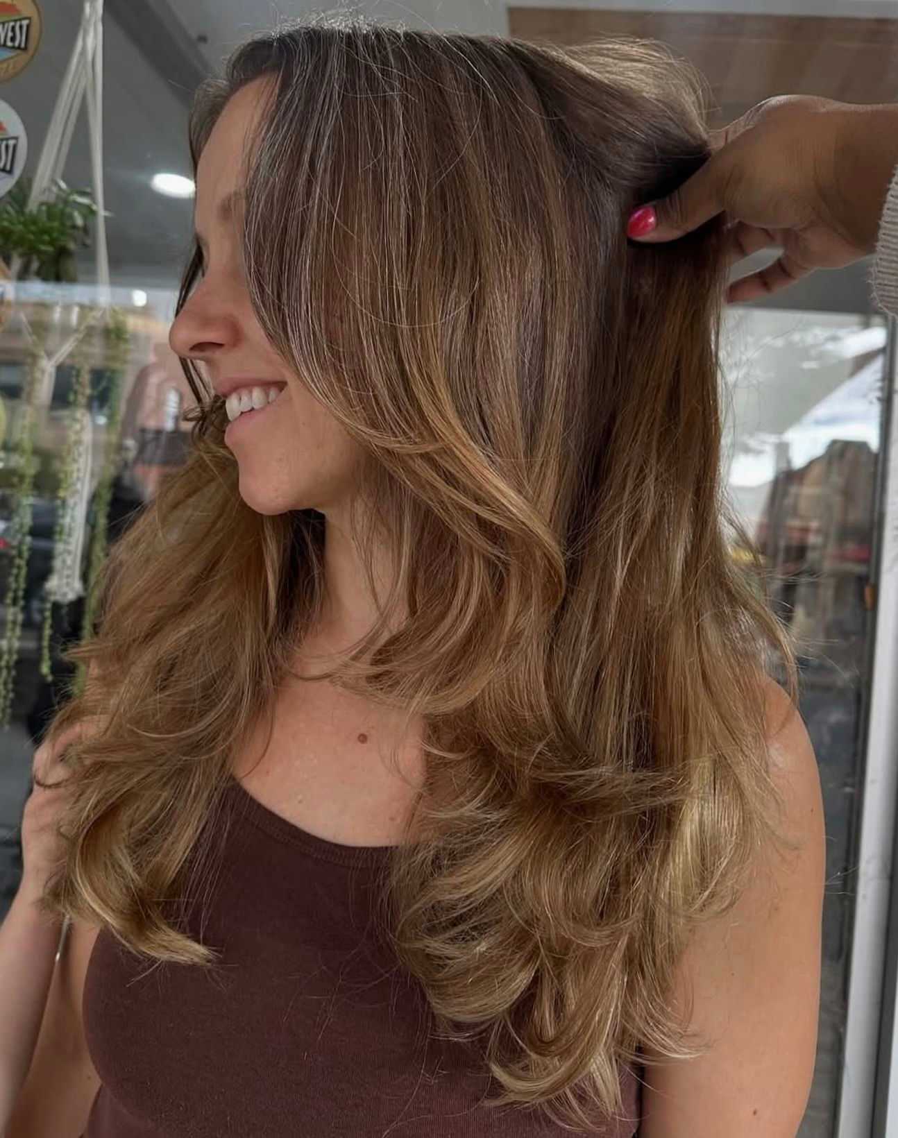 Woman with layered, wavy hair styled in salon, wearing a brown top.