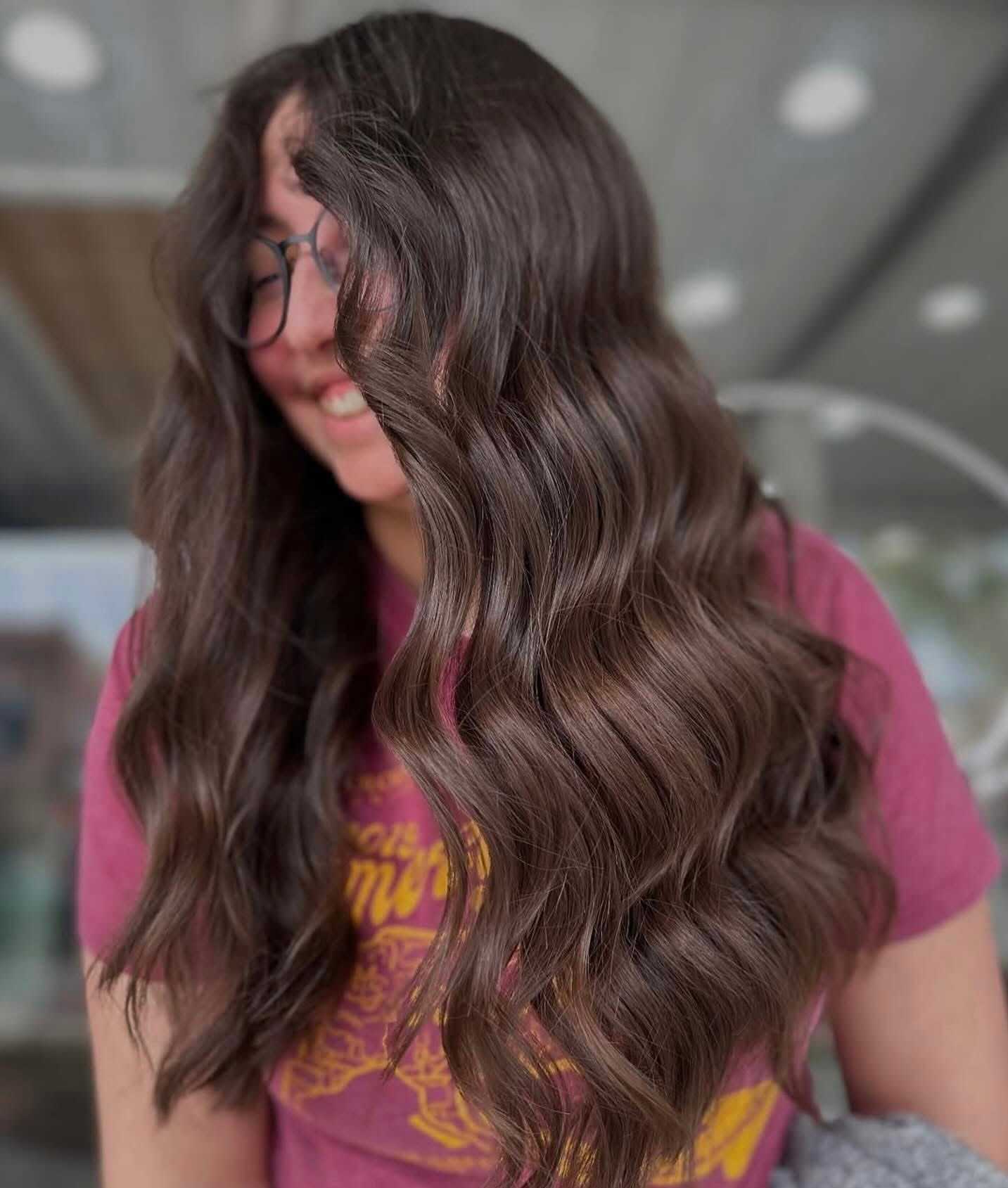 Woman with long, wavy hair and glasses, smiling in a casual setting.