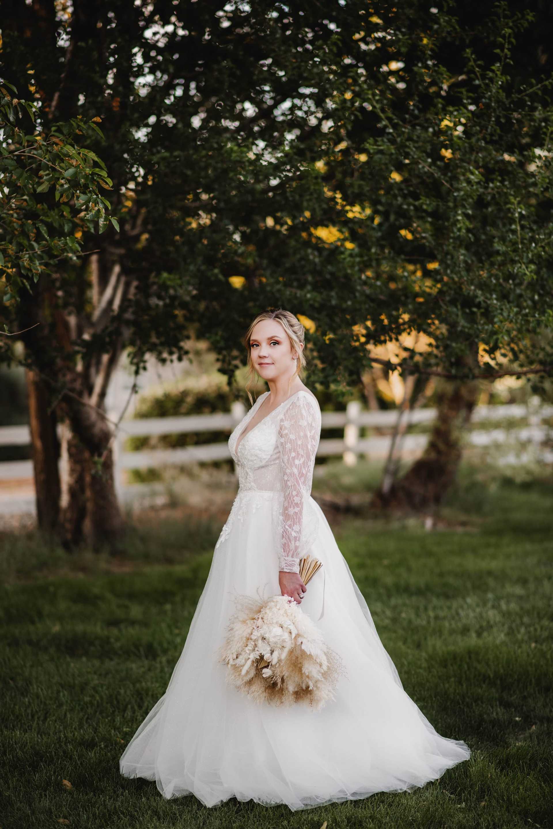 Bride in a white gown holding a bouquet, standing on grass with trees in the background.